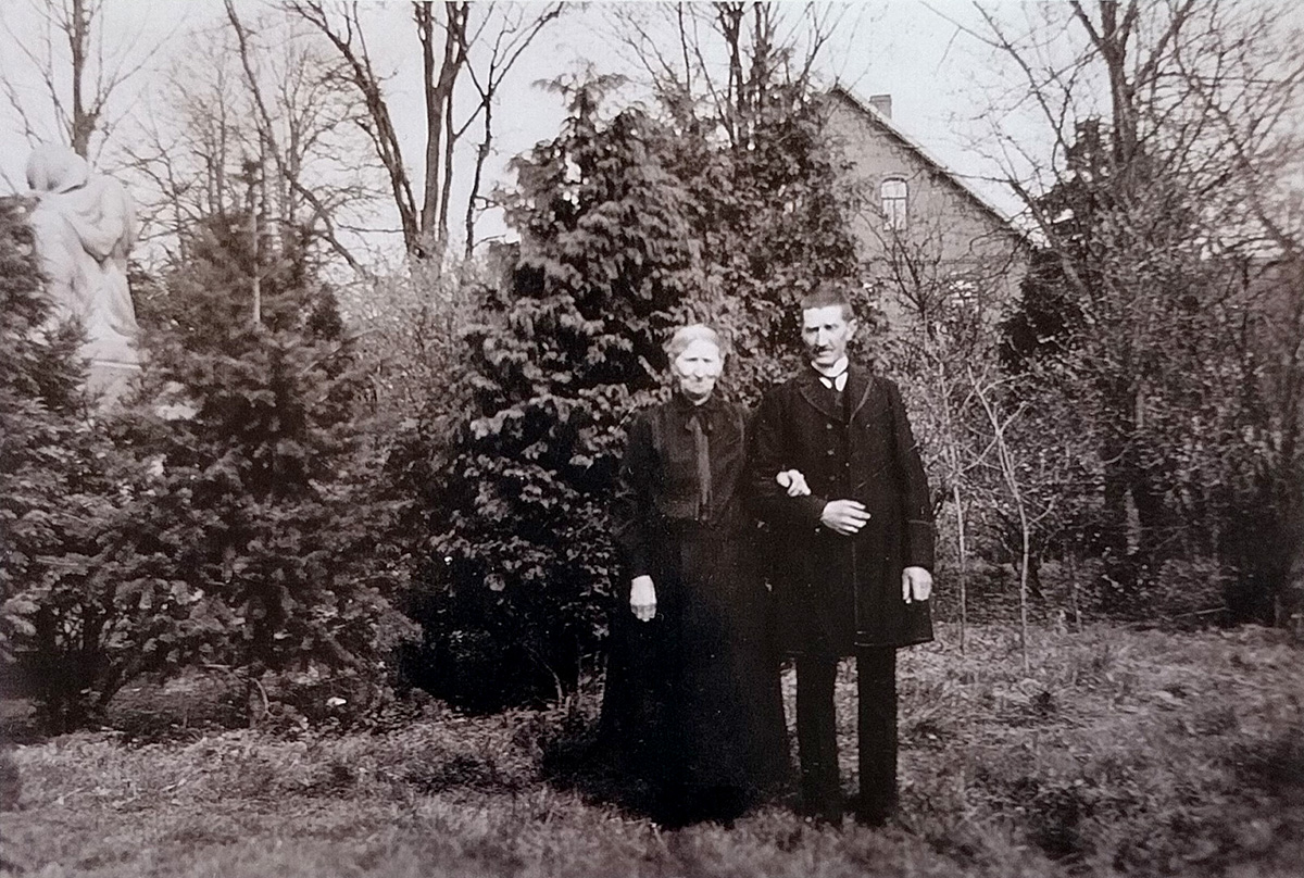 Es ist ein schwarz-weißes Foto. Es ist unscharf. Dora und Gustav Daps stehen auf einer Grasfläche vor einigen Bäumen. Dahinter ist ein Haus zu sehen. Dora trägt ein langes, hochgeschlossenes schwarzes Kleid. Ihre hellen Haare sind vermutlich grau und im Nacken zu einem Knoten zusammengebunden. Gustav trägt eine dunkle Hose und einen dunklen Mantel. Seine Haare sind kurzgeschnitten. Dora hat sich bei ihm untergehakt. Beide scheinen ernst in die Kamera zu blicken. Dora lächelt möglicherweise.