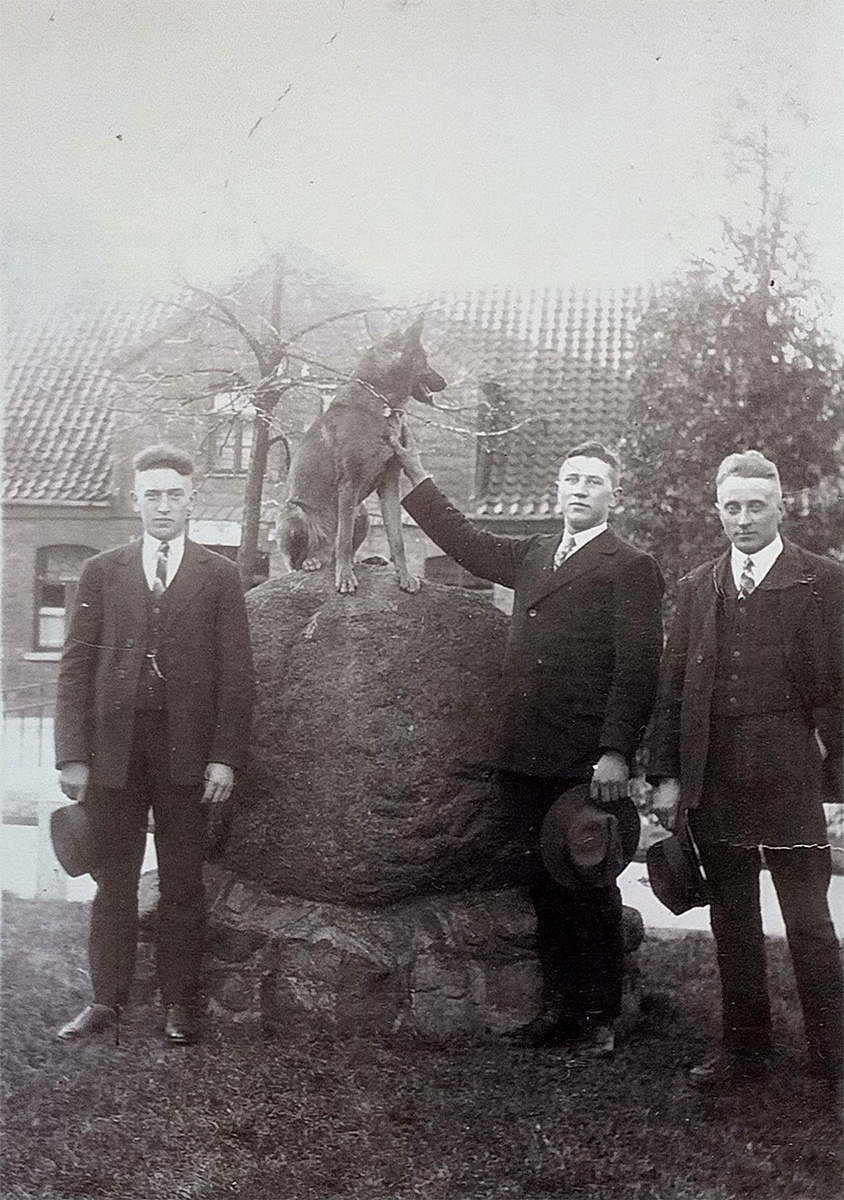 Es ist ein schwarz-weißes Foto. Drei junge Männer stehen neben einem großen Findling, der auf einem niedrigen Steinsockel liegt. Einer der Männer streichelt einen Schäferhund, der auf dem Findling sitzt. Die Männer sind fein gekleidet mit Anzug, Hemd und Krawatte. Sie halten ihre Hüte in den Händen und blicken in die Kamera. Im Hintergrund ist ein Haus.