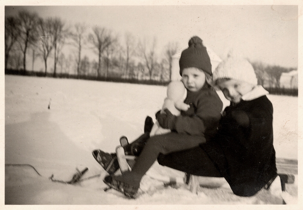 Es ist ein schwarz-weißes Foto. Ulla und Günter Schulze sitzen auf einem Schlitten und wurden von der Seite fotografiert. Ulla hält eine Puppe im Arm. Sie sitzt auf dem Schoß von Günter. Beide gucken zur Kamera.