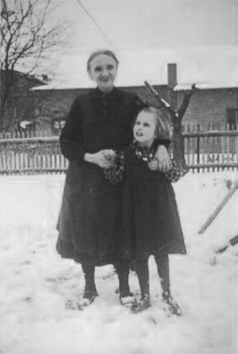 Das Foto ist sepia und leicht verschwommen. Christa steht vor ihrer Großmutter und wird von dieser an beiden Händen gehalten. Beide tragen dunkle langärmelige Kittelkleider. Und dunkle Strümpfe. Sie stehen im Schnee. Im Hintergrund sind landwirtschaftliche Gebäude zu erkennen.
