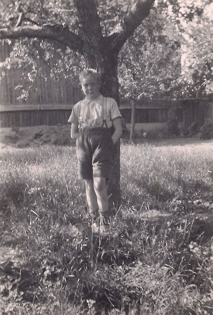 Das ist ein schwarz-weißes Foto. Heinz Knorr steht in einem Garten an einem Baumstamm. Er trägt eine kurze Hose mit Trägern. Dazu ein kurzärmeliges Hemd. Er blickt mit ernstem Gesicht zur Kamera.