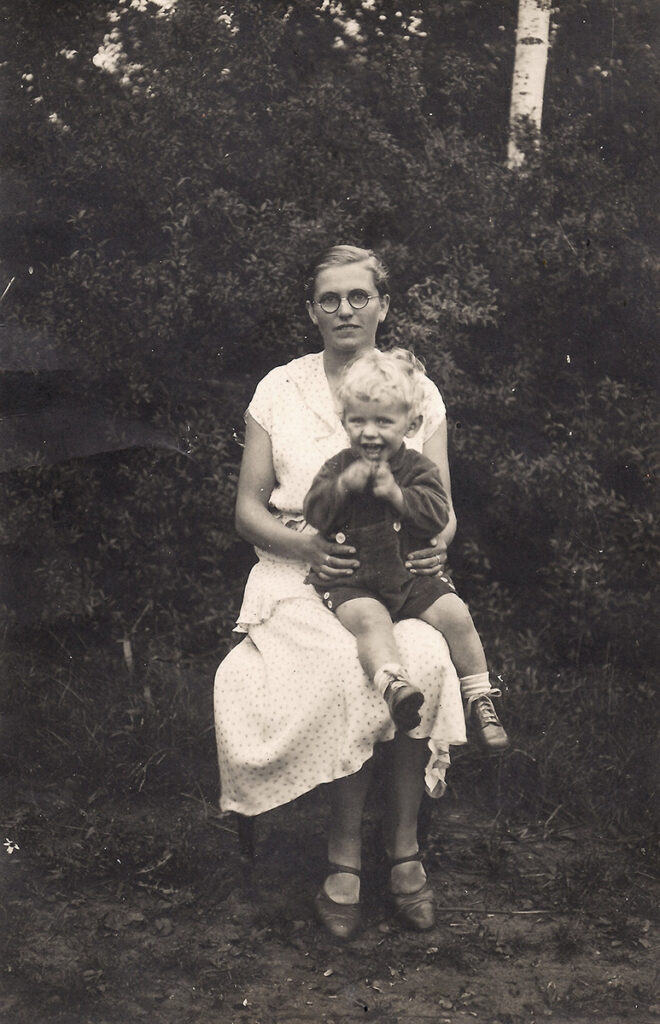 Es ist ein schwarz-weißes Foto von Rudolf auf dem Schoß seiner Mutter. Sie sitzen in einem Garten. Die Mutter trägt ein helles klein gemustertes Kleid. Ihr Sohn trägt eine kurze Latzhose und einen dunklen Pullover. Er hat blonde Locken und guckt sehr fröhlich. Er scheint gerade seine Hände zu bewegen. Seine Mutter trägt eine Brille und blickt lächelnd zur Kamera.