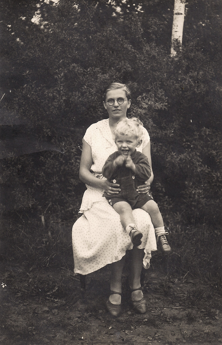 Es ist ein schwarz-weißes Foto von Rudolf auf dem Schoß seiner Mutter. Sie sitzen in einem Garten. Die Mutter trägt ein helles klein gemustertes Kleid. Ihr Sohn trägt eine kurze Latzhose und einen dunklen Pullover. Er hat blonde Locken und guckt sehr fröhlich. Er scheint gerade seine Hände zu bewegen. Seine Mutter trägt eine Brille und blickt lächelnd zur Kamera.