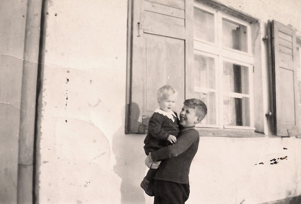 Es ist ein schwarz-weißes Foto. Die Geschwister Rudolf und Ingrid Hagedorn stehen vor einem Haus mit Fensterläden. Rudolf trägt dunkle Kleidung und hält seine Schwester auf dem Arm. Seine Schwester ist etwa ein Jahr alt. Sie trägt ein dunkles Kleid mit hellem Kragen. Beide gucken sehr vergnügt.