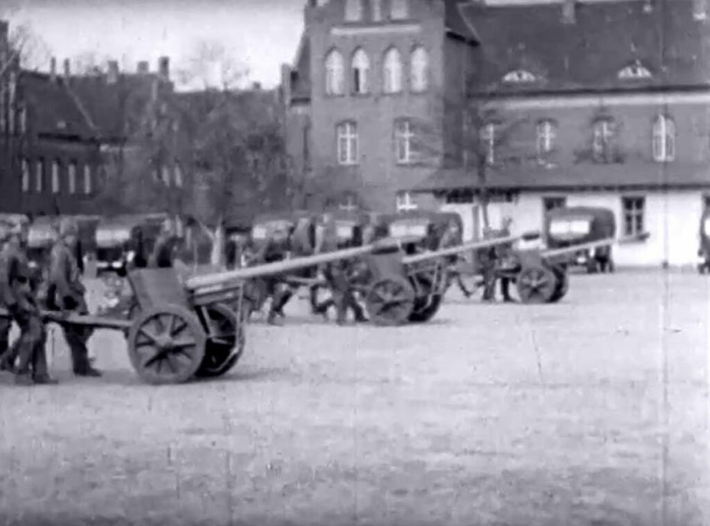 Es ist ein schwarz-weißes Foto. Es ist ein Standbild aus einem Film. Es zeigt einen Exerzierplatz mit Artillerie, die von deutschen Soltanten bedient wird. Im Hintergrund sind die Gebäude der ehemaligen Heil- und Pflegeanstalt Treskau-Owińsk zu erkennen.