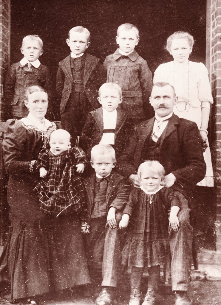 Es ist ein schwarz-weißes Foto. Die Familie hat sich in drei Reihen zum Foto aufgestellt. Die Eltern stehen mittig. Alle tragen festliche Kleidung. Alle blicken ernst in die Kamera.
