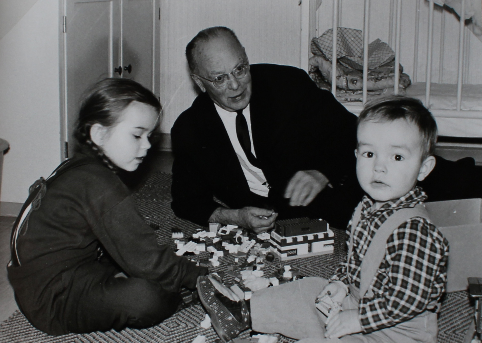 Es ist ein schwarz-weißes Bild. Zu sehen ist Heinz Heinze halb liegend am Boden mit zwei jungen Enkelkindern. Zwischen den dreien liegt ein Legohaus, an dem Heinz Heinze gerade zu bauen scheint. Er trägt einen dunklen Anzug mit Krawatte und wirkt gelöst.