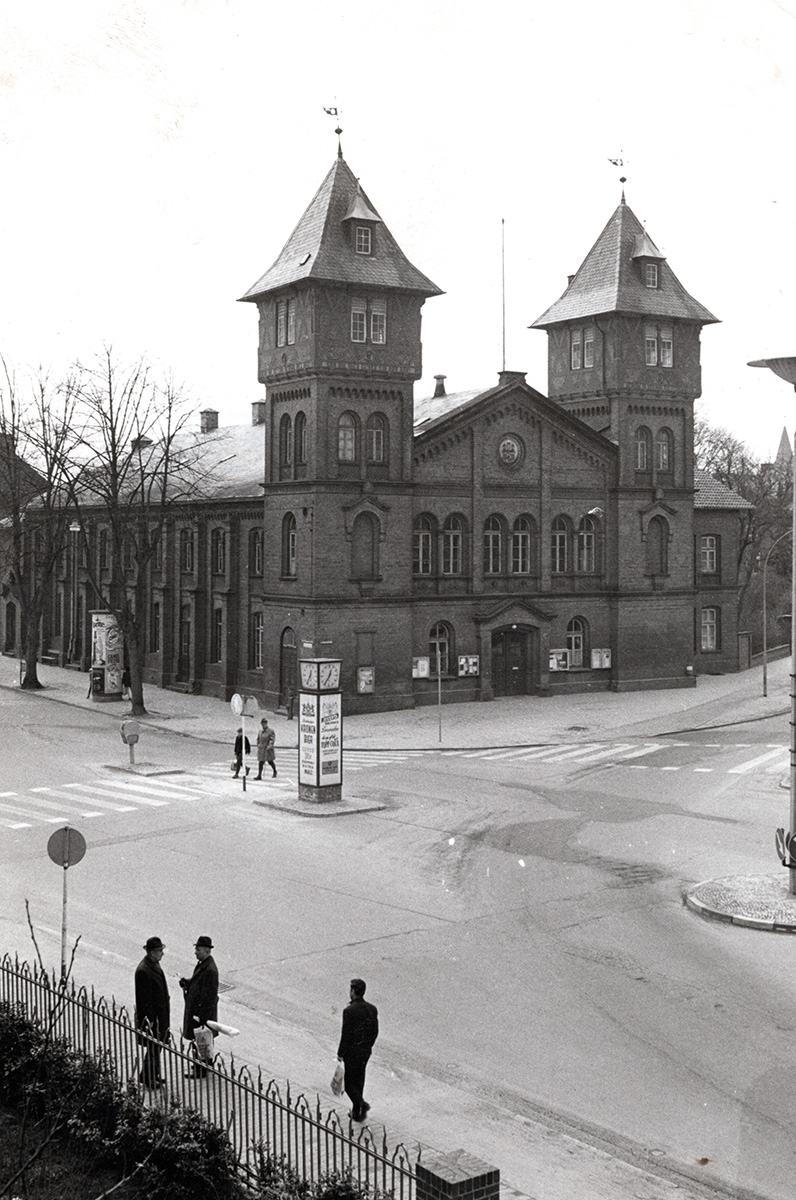 Es ist ein schwarz-weißes Bild. Aus einer erhöhten Position aufgenommen zeigt es eine Straßenkreuzung. Auf der gegenüberliegenden Seite steht die Turnhalle mit zwei markanten eckigen Türmen. Sie ist prachtvoll mit Backstein gemauert.