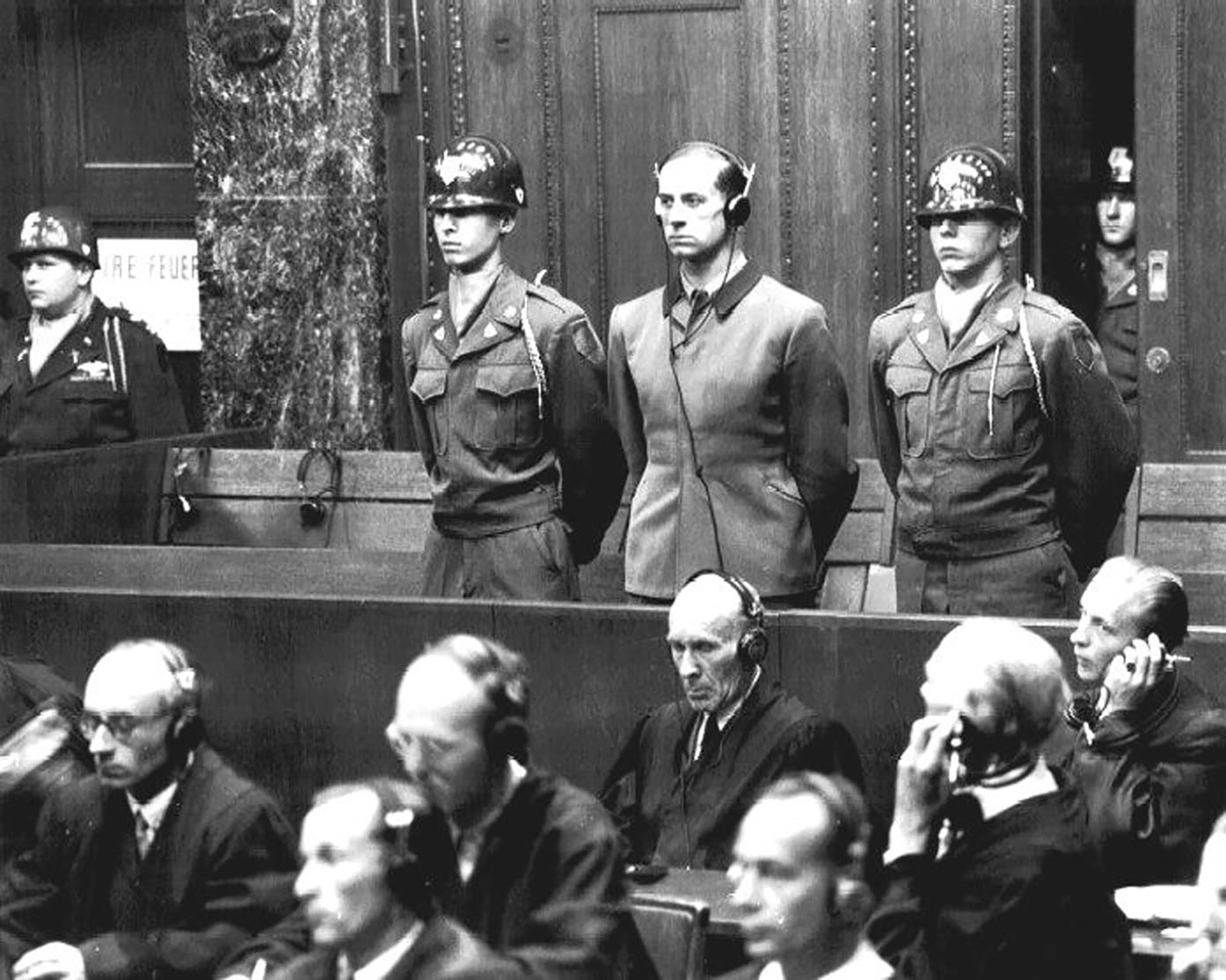 Es ist ein Schwarz-Weiß-Bild. Es zeigt den Blick auf den Angeklagten, der sich erhoben hat. Neben ihm steht auf jeder Seite ein uniformierter Militärpolizist. Brandt blickt ernst und trägt Kopfhörer mit Kabel. Die vor ihm sitzenden Juristen wirken vertieft in das, was sie ebenfalls über Kopfhörer hören. Nur einer von ihnen dreht sich zu ihm um. Im Hintergrund sind die Marmorsäulen und schweren Holzvertäfelungen des Raumes zu erkennen.