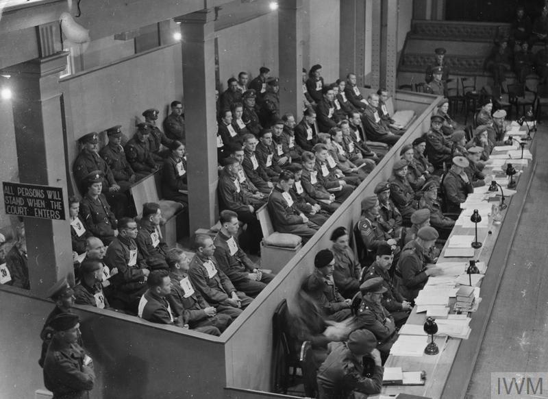Es ist ein schwarz-weißes Foto. Zu sehen ist ein Blick von oben auf eine große Menge sitzender Mesnchen. Alle sind zum rechten Bildrand hin ausgerichtet. Die ersten zwei Reihen bilden Menschen mit militärischer Kleidung an einem langen Tisch mit Papieren und Leselampen. Dahinter sitzen abgetrennt durch eine Brüstung drei Reihen Menschen mit einheitlicher Kleidung und Nummern vorne auf der Brust. Hinter ihnen sitzen Personen in Uniform.