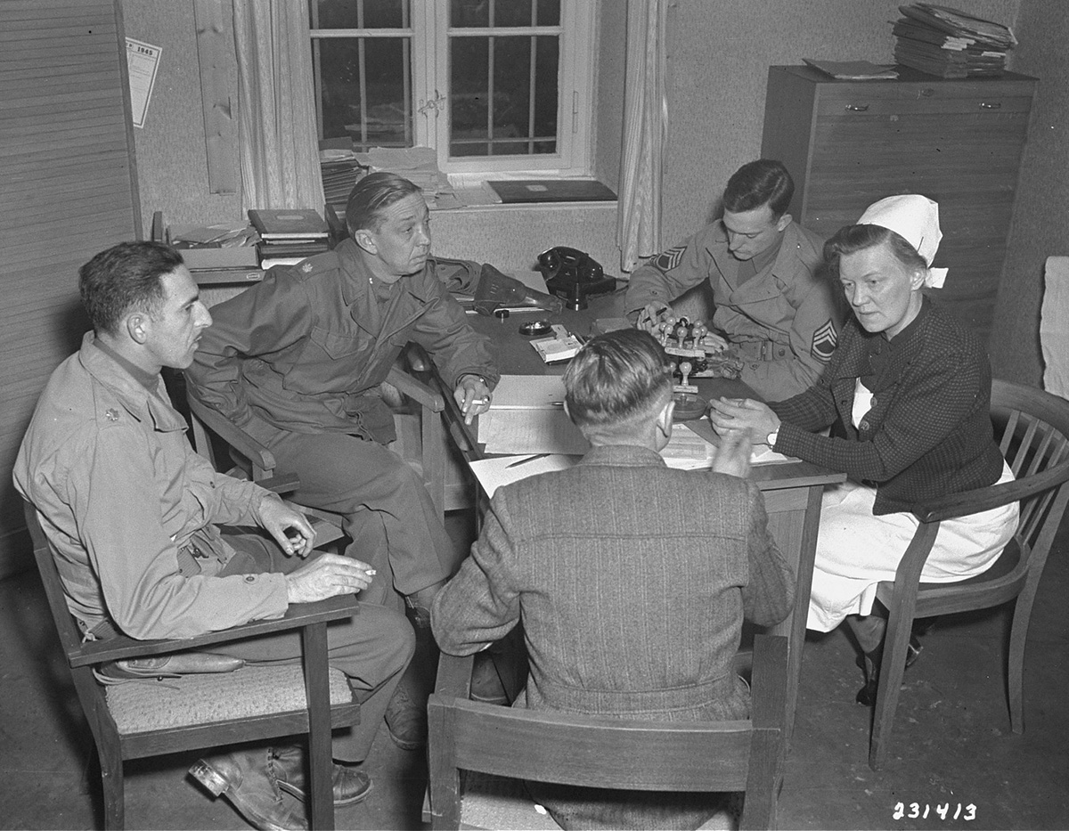 Es ist ein schwarz-weißes Foto. In einem kleinen Raum befinden sich vier Männer und eine Frau. Sie sitzen um einen Holztisch. Die Männer tragen Uniform, die Frau Schwesternkleidung. Auf dem Tisch liegen einige Papiere und Stempel.