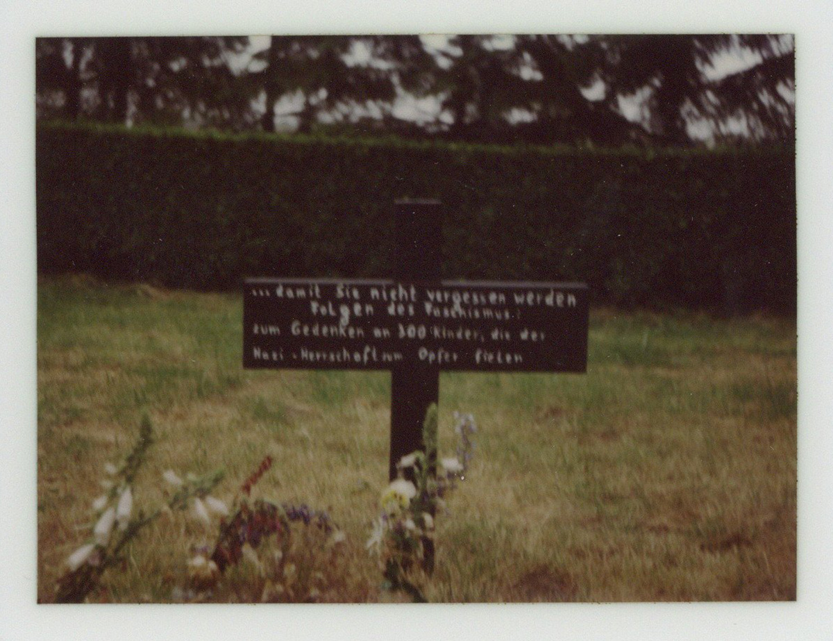 Es ist ein verschwommenes Farbfoto. Es zeigt zentral ein niedriges Holzkreuz mit Inschrift. Vor dem Kreuz stehen drei schmale Blumensträuße.