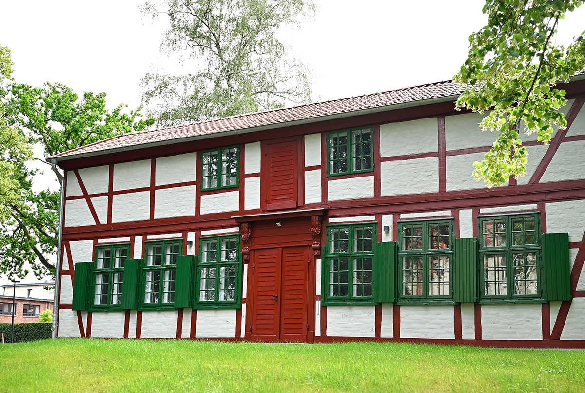 Es ist ein Farbfoto. Darauf ist ein niedriges zweistöckiges Fachwerkhaus. Es hat weiße Wände, rote Balken und dunkelgrüne Fenster mit Fensterläden.
