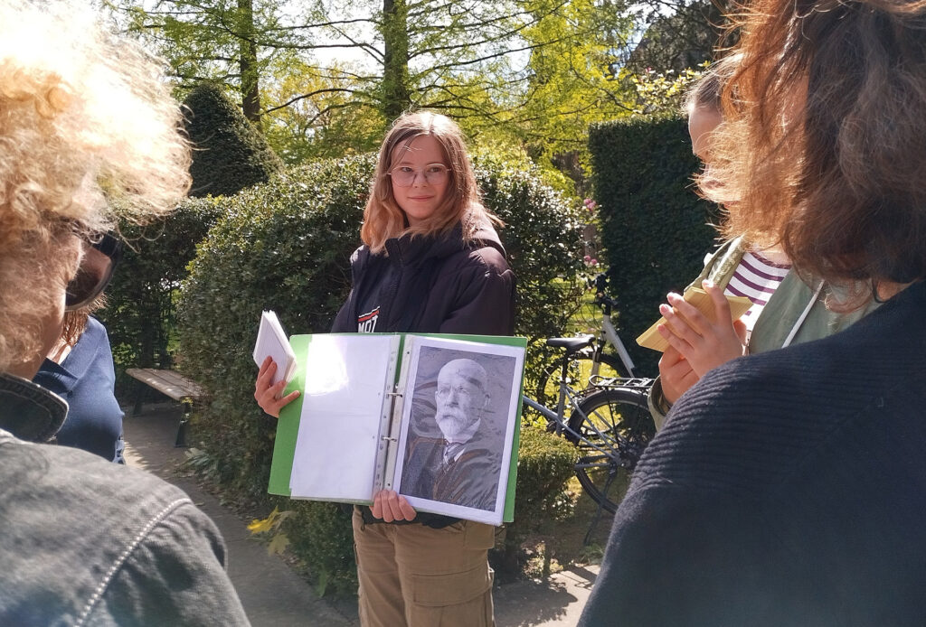 Es ist ein Farbfoto. Zwischen zwei Personen hindurch wurde eine Schülerin fotografiert. Diese präsentiert in einer Mappe ein Foto von Otto Snell. Die Schülerin guckt freundlich die umstehenden Personen an.