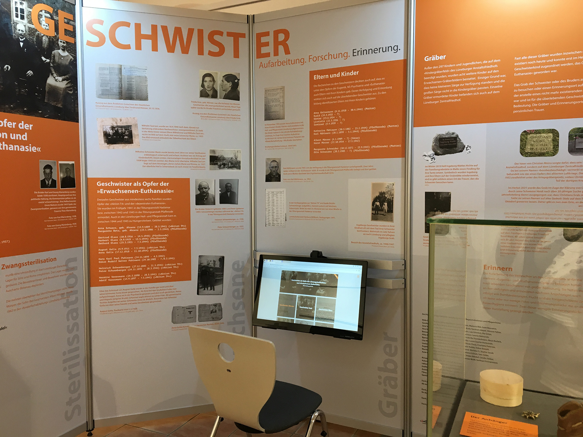Es ist ein Farbfoto. Große Posterwände der Ausstellung in weiß-oranger Farbgestaltung sind zu sehen. Vor einer Wand ist ein Bildschirm, vor dem ein Stuhl bereitsteht.
