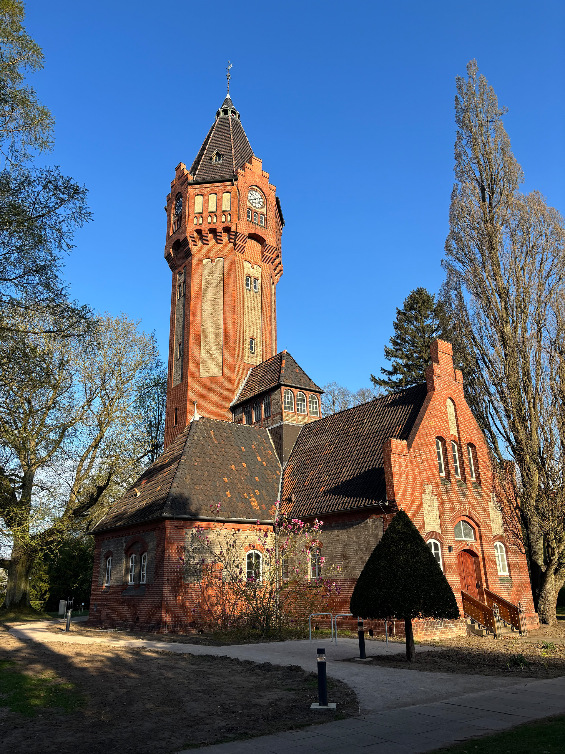 Es ist ein Farbfoto. Es zeigt den Wasserturm von unten fotografiert. Sonnenschein lässt die farbigen Backsteine leuchten. Der Himmel ist blau.
