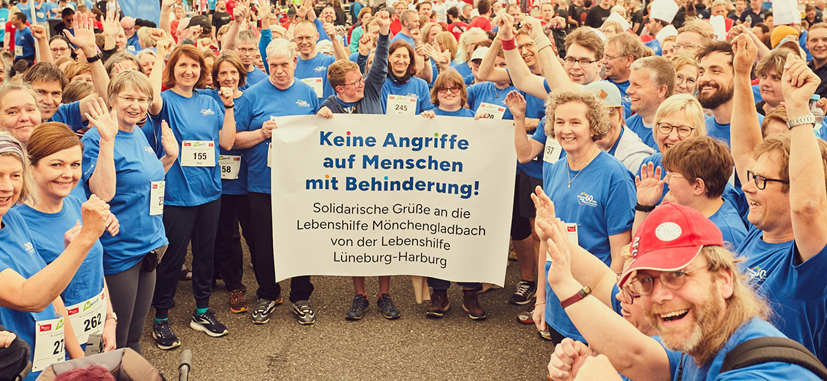 Eine große Gruppe Menschen in blauen T-Shirts steht um ein Transparent herum. Sie blicken kämpferisch zur Kamera.