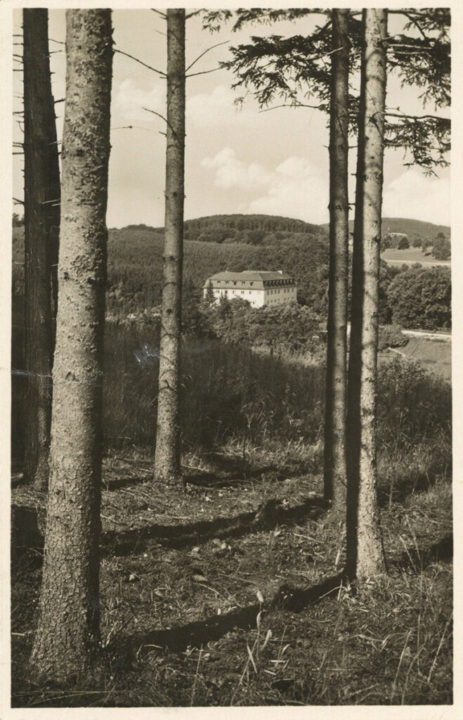 It is a black and white picture. It shows the view of Grafeneck. There are several tall tree trunks in the foreground. The hunting lodge can be seen between the trunks. It is an angular white building with a dark roof. There are many windows on all sides. It is surrounded by dense forest on a hill.
