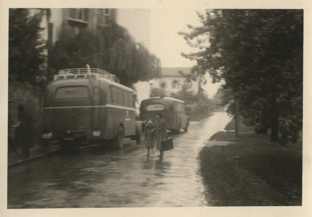 Es ist ein Schwarz-Weiß-Bild. Es zeigt eine Straße vor dem Jagdschloss. Auf der Straße stehen am linken Rand vor dem Gebäude zwei alte Reisebusse. Neben den Bussen sind zwei Personen. Eine größere Person mit Koffer in der Hand hält eine kleinere Person am Arm. Es scheint zu regnen. Die Personen tragen Mäntel.