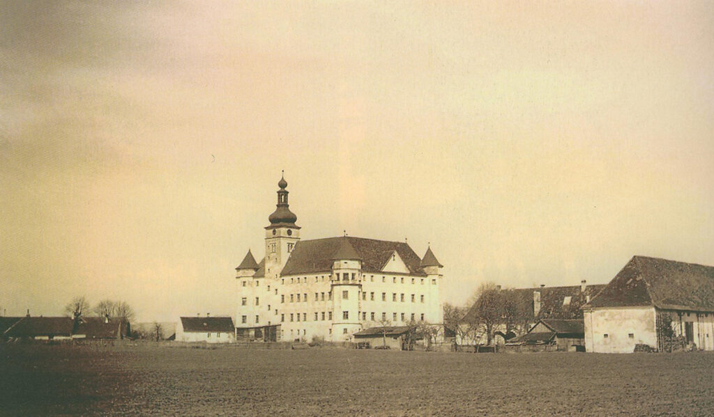 Es ist ein Schwarz-Weiß-Bild. Es ist leicht vergilbt. Inmitten von Feldern liegt ein helles viertstöckiges Gebäude mit einem hohen zwiebelförmigen Glockenturm. Das Gebäude hat viele Fenster in jedem Stockwerk und ein hohen dunkles Dach. An den Ecken sind kleine runde Erker mit Turm obenauf.
