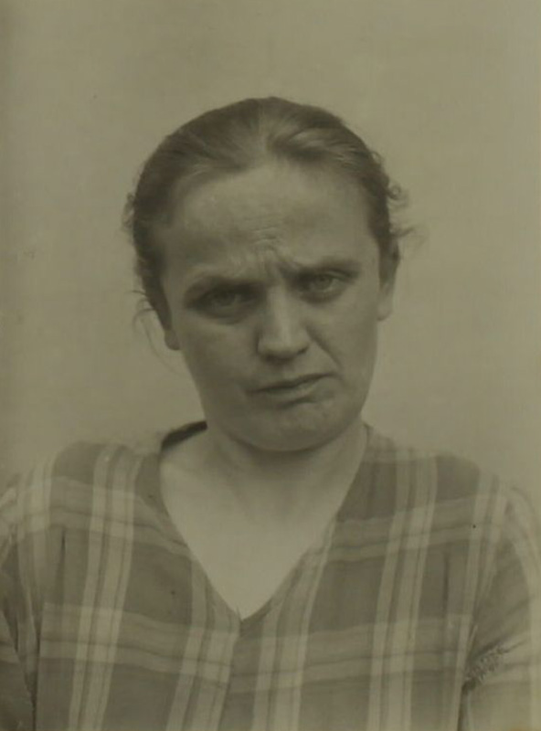 Es ist ein schwarz-weißes Foto. Es ist ein Porträt von Olga Schulze. Sie ist eine junge Frau und trägt ein kariertes Oberteil. Ihre Haare sind zu einem Zopf nach hinten gebunden. Sie blickt ernst und verstört in die Kamera.