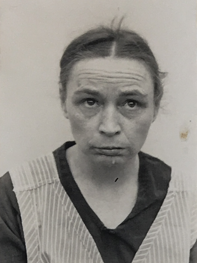 Es ist ein schwarz-weißes Porträtfoto. Betty Fleck trägt einen gestreiften Kittel über einer dunklen Bluse. Ihr Haar ist nach hinten gebunden, einige Strähnen stehen heraus. Sie hat die Stirn in Falten gelegt und guckt nach oben.