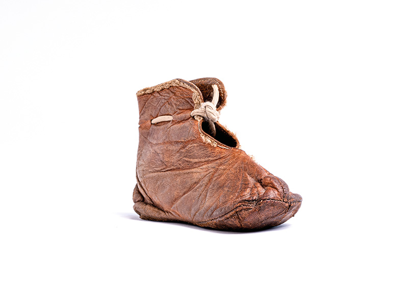 Das ist ein Farbfoto von eine, Kinderschuh. Der Schuh ist aus braunem Leder. Er hat eine Stiefelform. Das Schnürband führt oben zusammen.