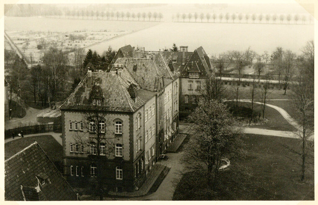 Es ist ein schwarz-weißes Bild. Die Postkarte ist vergilbt. Aus einer erhöhten Position zeigt das Bild einen Blick auf die Heil- und Pflegeanstalt. Es sind zwei dreistöckige, helle Gebäude mit dunklem Dach zu sehen. Die Gebäude sind von einem angelegten Garten umgeben.