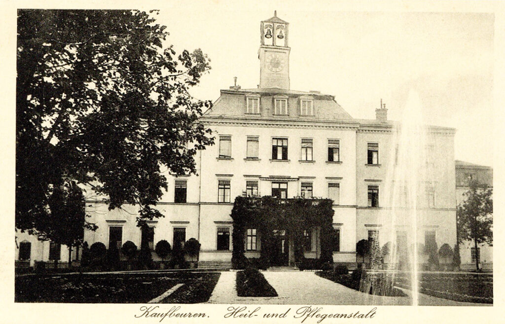 It is a black and white picture. A park with a fountain leads up to a stately-looking building. The building is light-coloured and has three floors. There is a bell tower on the roof above the entrance.