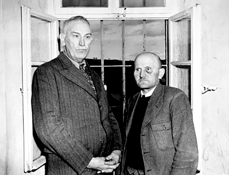 It is a black and white picture. Two men are standing in front of an open window with casements. The man on the left is taller. He is wearing a striped suit with a shirt and tie. The man on the right is smaller. He is wearing a coarse dark suit with a jumper and shirt. He appears to have a black eye. Both of them have their hair slicked back. They are facing each other but look past each other out of the picture.