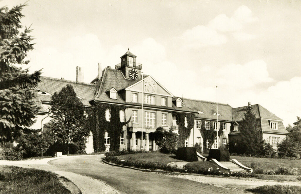 It is a black and white picture of the main house. A landscaped park can be seen in front of the house. The house has an entrance portal and a wing on each side. Above the entrance is a large clock with a bell tower.