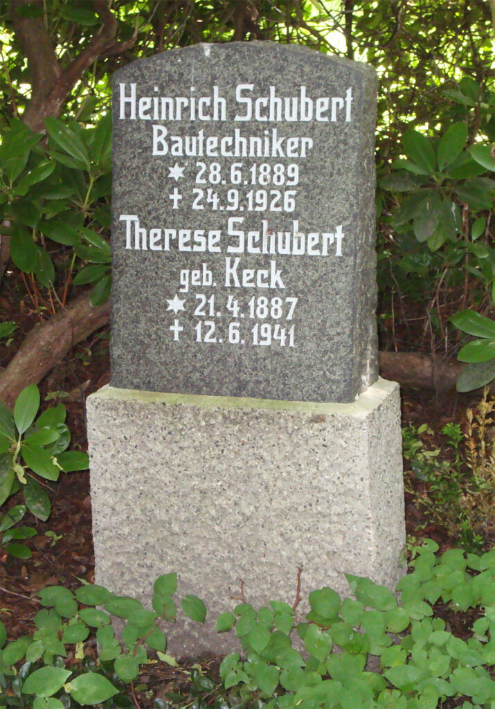 Es ist ein Farbfoto von einem Grab. Der Grabstein ist aus grauem Granit. Auf einem Sockel ist ein polierter Stein mit eingravierten Namen und Lebensdaten. Der Stein ist oben oval abgeschlossen. Der Grabstein ist umgeben von grünen Sträuchern.