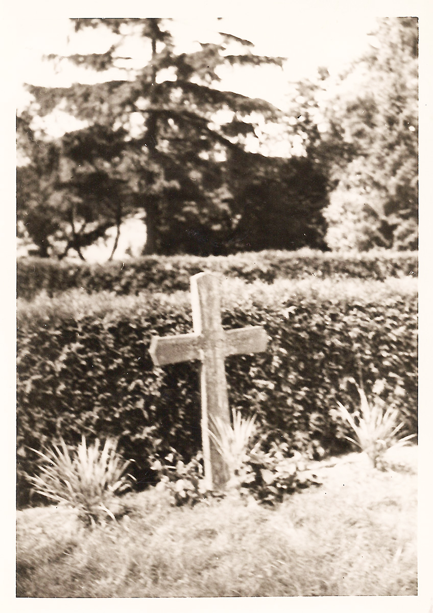 Es ist ein schwarz-weißes Foto von einem Einzelgrab. Zu sehen ist ein Holzkreuz mit einer eingravierten Grabnummer. Hinter dem Kreuz verläuft eine Hecke. Das Kreuz ist von einer schlichten Rasenbepflanzung eingefasst.