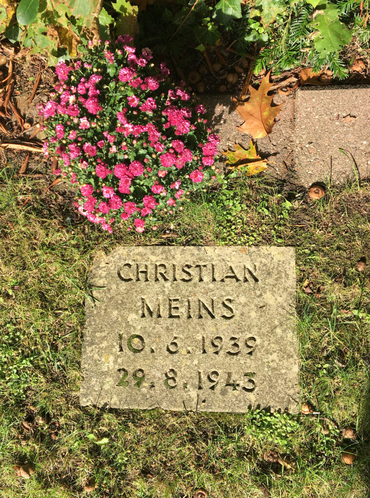 Es ist ein Farbfoto vom Grab von Christian Meins. Es ist ein Kissenstein. Auf dem Stein steht: Christian Meins, 10. Juni 1939, 29. August 1943. Der Kissenstein ist von Rasen umgeben. Oberhalb des Steins steht ein Topf mit rosafarbenen Blumen. Der Topf steht auf einer Rasenkante, die den Rasen oberhalb des Grabsteins abschließt.