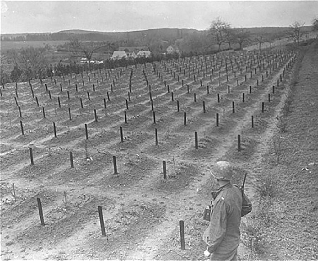 Es ist ein schwarz-weißes Foto. Es ist von leicht erhöht aufgenommen. Sichtbar ist ein weites, leicht abschüssiges Gräberfeld. Auf dem Gräberfeld markieren symmetrisch angeordnete Holzpflöcke die einzelnen Gräber. Jeder Holzpflock trägt eine Grabnummer. Ein Soldat in amerikanischer Uniform steht mit Helm und Gewehr am Rand des Gräberfeldes und blickt in Richtung der Gräber.