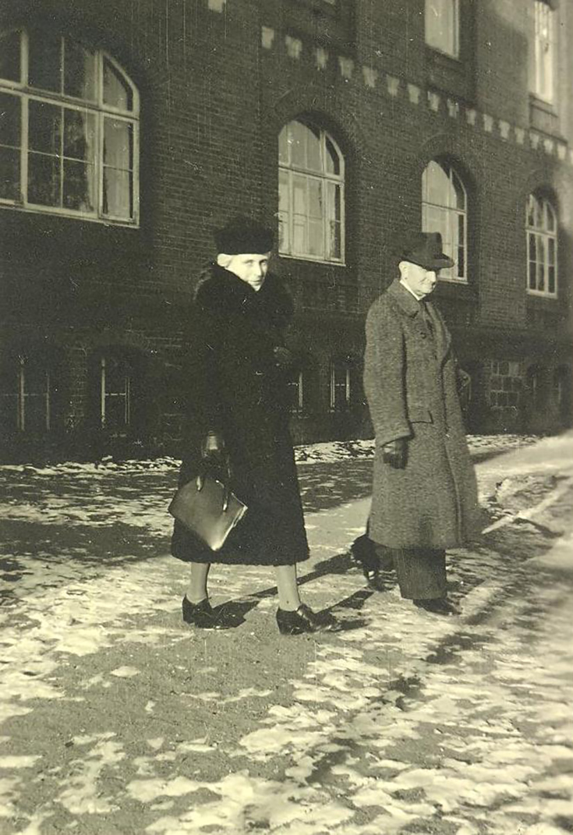 Es ist ein schwarz-weißes Foto des Ehepaars Emililie und Gustav Marx. Emilie trägt einen schwarzen Pelzmantel und einen Pelzhut, Gustav Marx trägt einen Hut, einen langen Wollmantel und Lederhandschuhe. Sie laufen über den gepflasterten Weg, der vor dem Verwaltungsgebäude der Anstalt verläuft. Sie Szene wirkt als würden sie festlich gekleidet zu einer Feier aufbrechen, das Foto ist ein Schnappschuss. Beide blicken spontan in die Kamera.